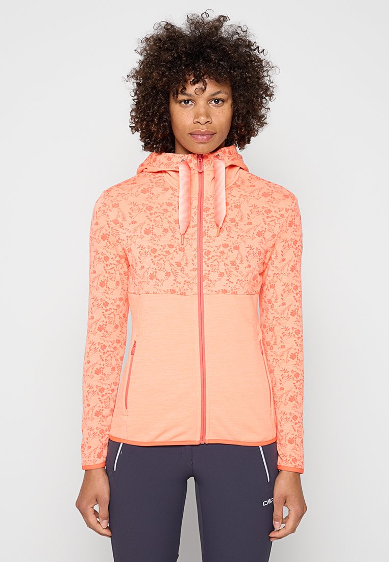 Frau mit lockigem Haar, die einen pfirsichfarbenen, mit Blumen bedruckten Zip-Hoodie und dunkle Sportleggings trägt, stehend vor einem einfarbigen hellen Hintergrund.