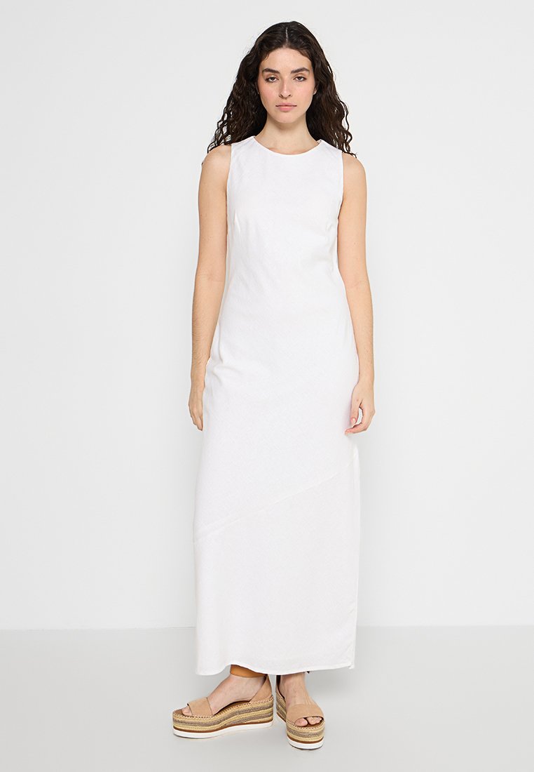 Weißes ärmelloses Maxikleid aus strukturiertem Stoff, mit einem taillierten Oberteil und einem asymmetrischen Saum. Kombiniert mit Plateausandalen.