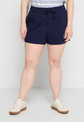 Person trägt marineblaue Shorts mit Kordelzug, beige Sneaker und ein gestreiftes, in die Hose gestecktes Hemd, steht vor einem schlichten hellen Hintergrund.