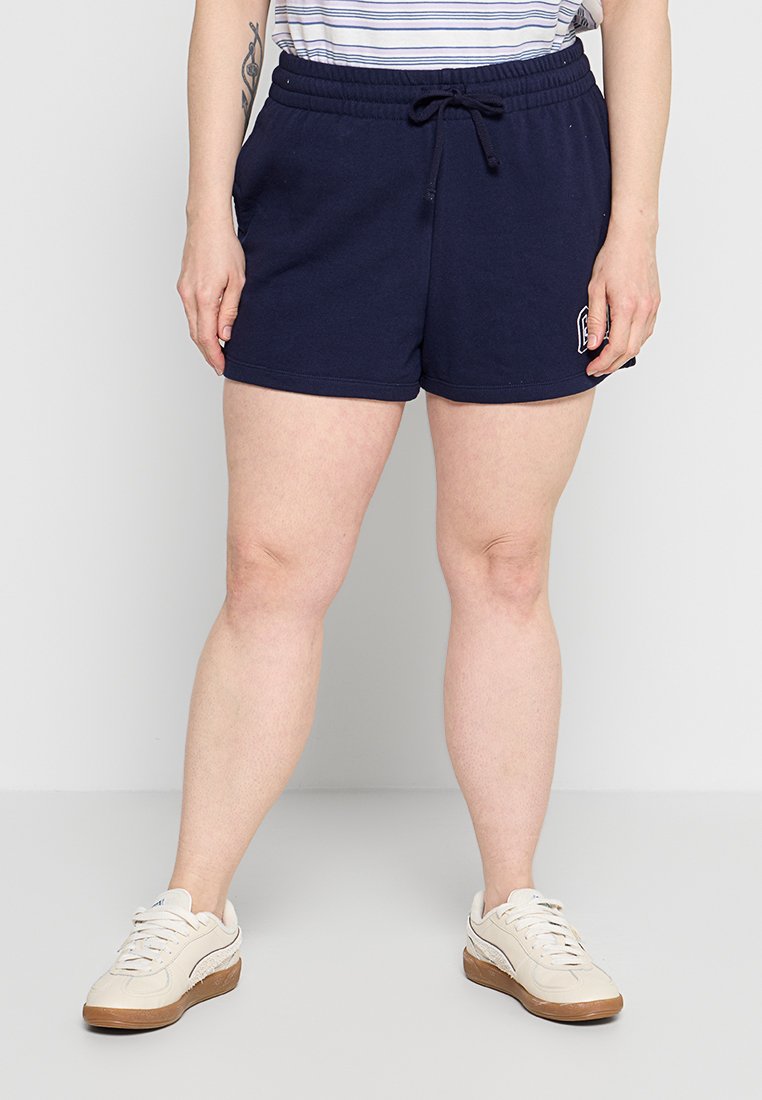 Person trägt marineblaue Shorts mit Kordelzug, beige Sneaker und ein gestreiftes, in die Hose gestecktes Hemd, steht vor einem schlichten hellen Hintergrund.