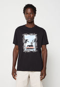 Mann trägt schwarzes T-Shirt mit Grafik einer Person, die auf einem Van steht, umgeben von Palmen vor einem Strand-Hintergrund.