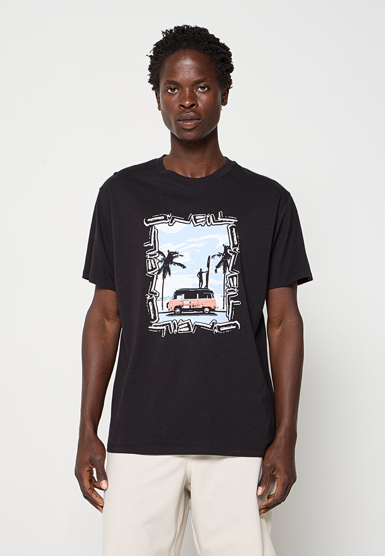 Mann trägt schwarzes T-Shirt mit Grafik einer Person, die auf einem Van steht, umgeben von Palmen vor einem Strand-Hintergrund.