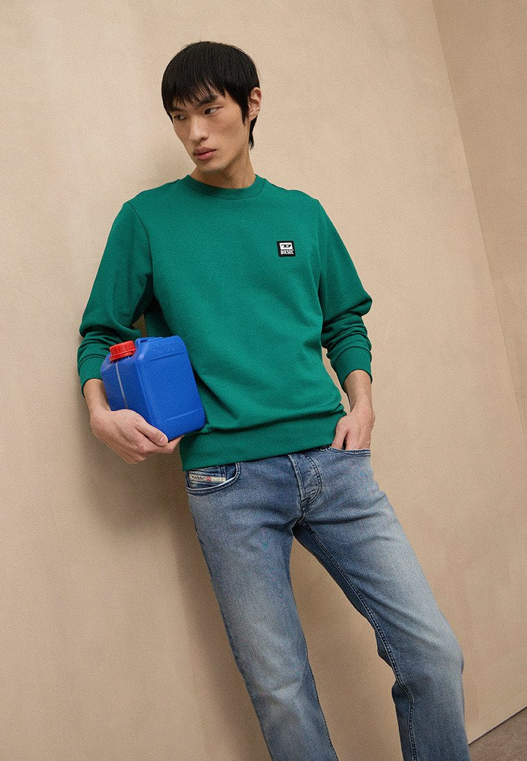 Grüner Sweatshirt mit Rundhalsausschnitt und kleinem Logoaufnäher, kombiniert mit hellblauen Jeans. Das Modell hält einen blauen Behälter mit rotem Deckel.