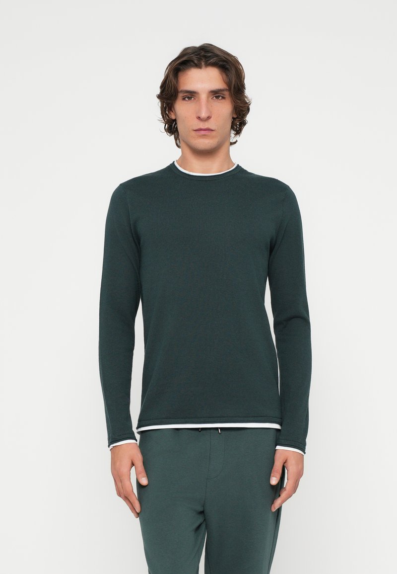 Dunkelgrünes Langarmshirt mit rundem Ausschnitt, versehen mit weißem Besatz am Kragen und an den Manschetten, gefertigt aus einem strukturierten Stoff.
