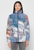 Frau, die eine Reißverschlussjacke mit hohem Kragen und einem abstrakten, mehrfarbigen Muster in Blau, Weiß und Orange trägt, steht vor einem schlichten Hintergrund.