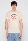 Junger Mann trägt beige T-Shirt mit rotem und schwarzem "Schott Motor Club" Text und Totenkopf-Design, kombiniert mit hellblauen Jeans, steht vor einfachem Hintergrund.