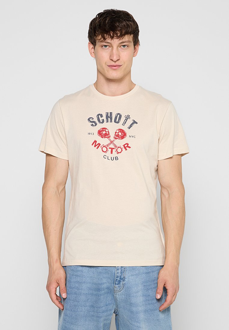 Junger Mann trägt beige T-Shirt mit rotem und schwarzem "Schott Motor Club" Text und Totenkopf-Design, kombiniert mit hellblauen Jeans, steht vor einfachem Hintergrund.