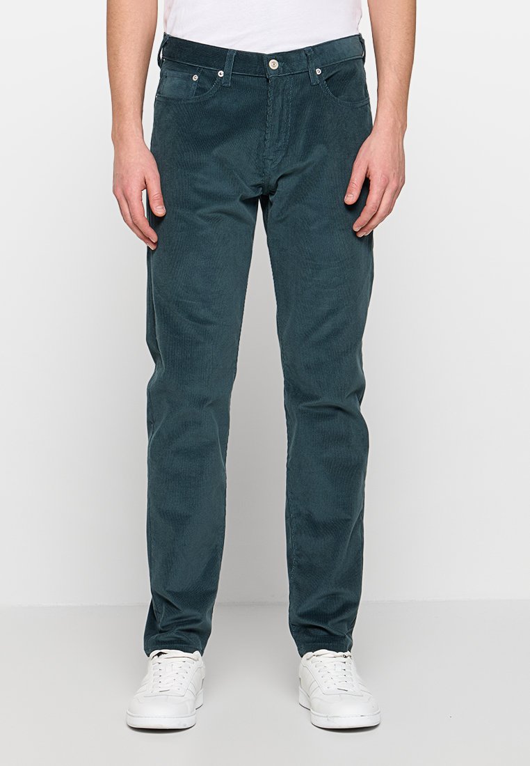 Tealfarbene Cordhose mit geradem Bein, ausgestattet mit Reißverschluss, fünf Taschen und subtiler Textur. Kombiniert mit weißen Turnschuhen.