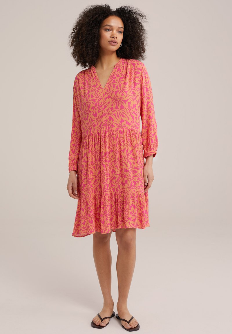 Rotes Kleid mit floralem Muster, V-Ausschnitt, langen Ärmeln und gestuftem Rock, aus leichtem Stoff gefertigt. Getragen mit schwarzen Ballerinas.