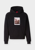 Schwarzer Kapuzenpullover mit Fronttasche und einem Sepia-Berg- und Baumgrafik zentriert über der Tasche mit rotem "HUGO"-Schriftzug.