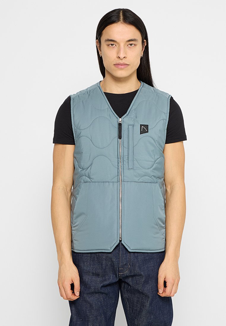 Person mit langen Haaren, die eine hellblaue gesteppten Weste über einem schwarzen Kurzarmshirt und dunklen Jeans trägt, steht vor einem weißen Hintergrund.