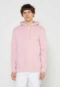 Junger Mann mit kurzem lockigem Haar, der einen hellrosa Kapuzenpullover und weiße Hosen trägt, vor einem einfarbigen hellen Hintergrund steht.