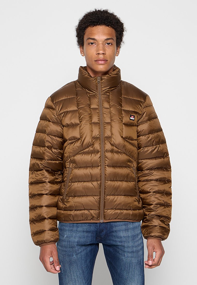 Braune Puffjacke mit hohem Kragen, gestepptem Design, Reißverschluss und Logo-Patch auf der Brust. Getragen über blauen Jeans.