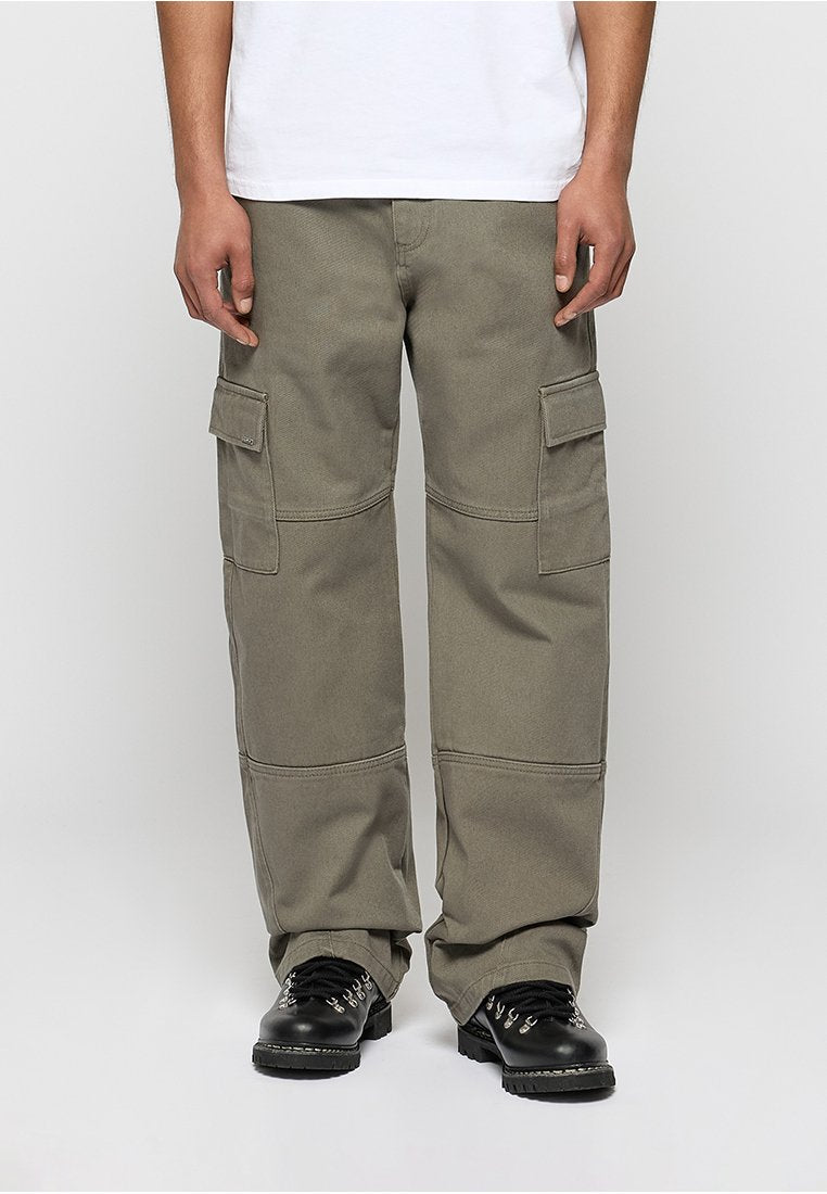 Mann trägt lockere olivgrüne Cargohosen mit Seitentaschen und schwarze Schnürstiefel, steht vor einem einfarbigen hellen Hintergrund.
