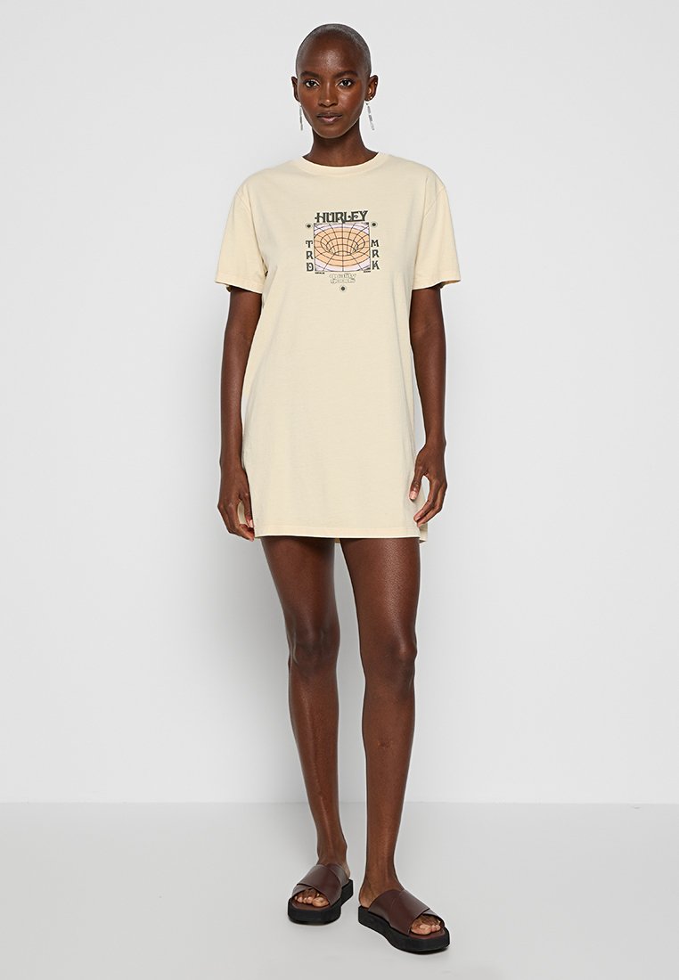 Cremfarbendes T-Shirt-Kleid mit kurzen Ärmeln und einem grafischen Design in der Mitte. Hergestellt aus weicher Baumwolle, hat es einen lockeren Schnitt und endet oberhalb der Knie.