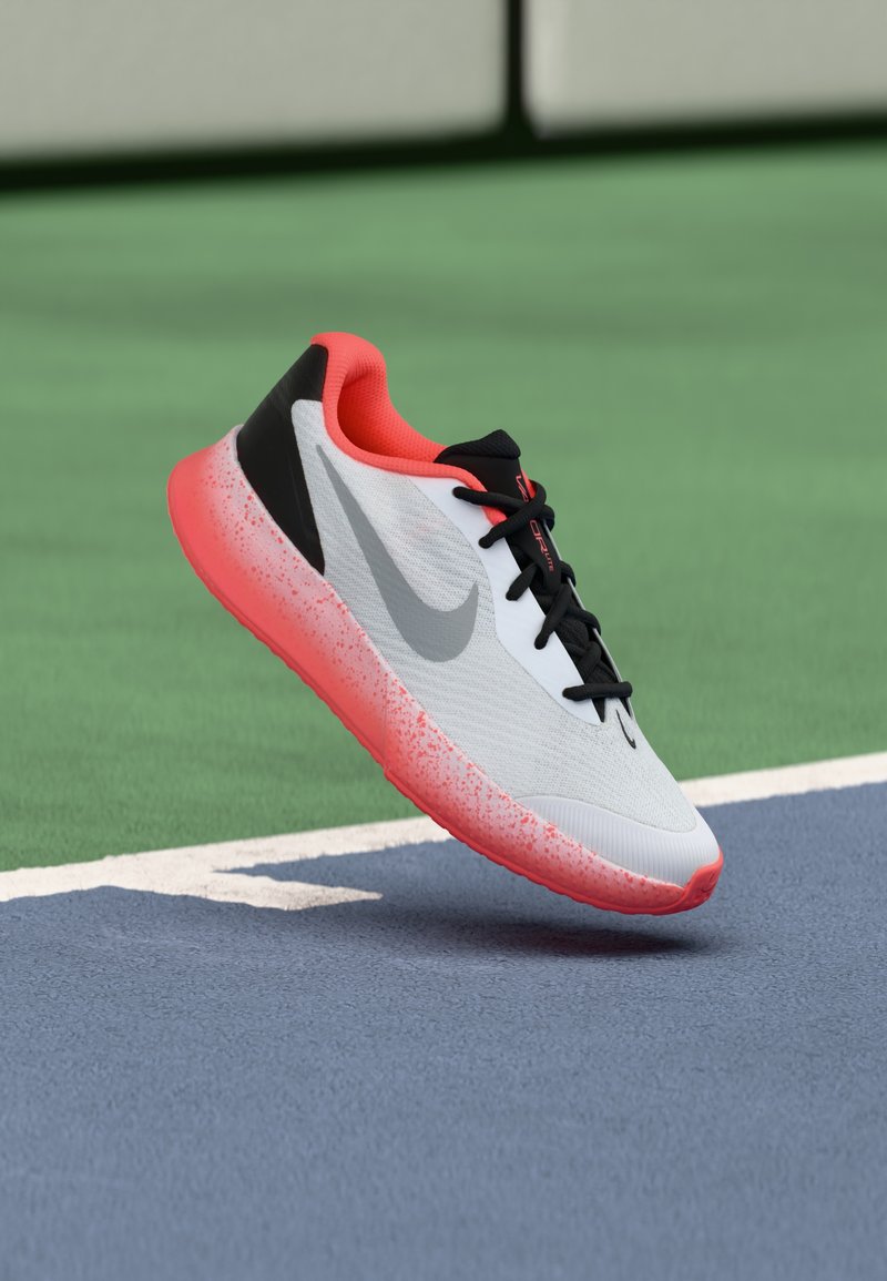 Weißer Sportschuh mit einer gemusterten, pinkfarbenen Gummisohle, schwarzen und korallenfarbenen Akzenten sowie einem strukturierten Obermaterial. Markantes graues Logo vorhanden.