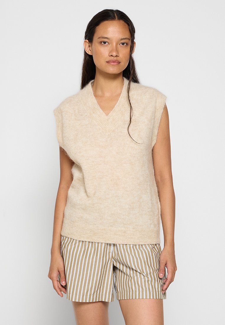 Beige gestrickter ärmelloser Pullover mit V-Ausschnitt, weicher Textur und lockerer Passform, kombiniert mit braunen gestreiften Shorts.