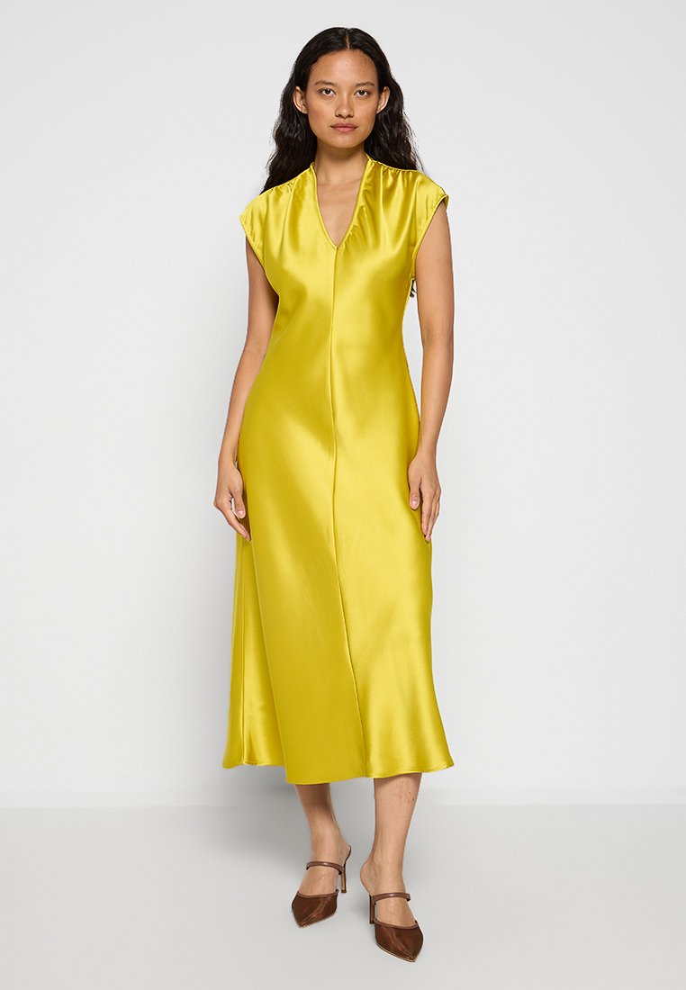 Gelbes Satin-Kleid mit kurzen Ärmeln, einem tiefen V-Ausschnitt und einer geraden Silhouette, die bis zur Mitte der Waden reicht. Trägt dazu braune Absatzschuhe.