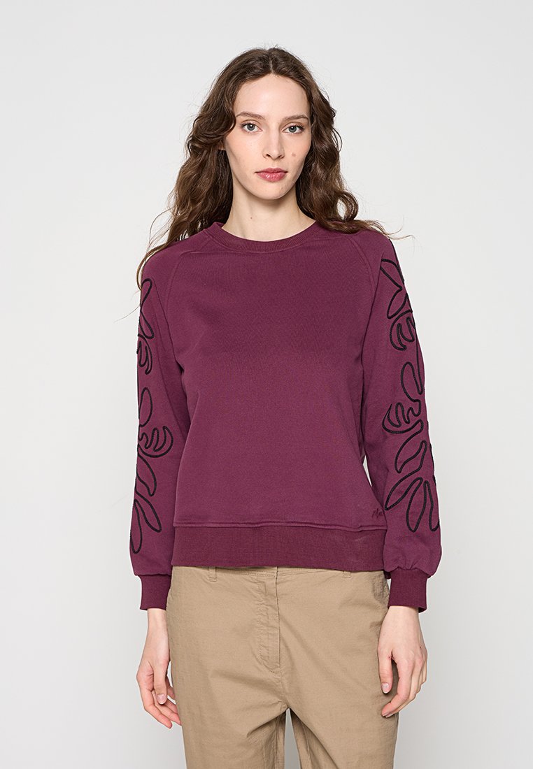 Frau mit langen braunen Haaren, die einen bordeauxroten Pullover mit schwarzen abstrakten Ärmeldesigns und beige Hosen trägt, steht vor einem einfarbigen Hintergrund.