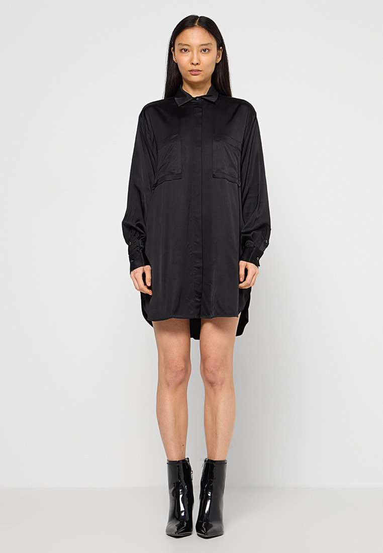 Schwarzes Satinhemdkleid mit langen Ärmeln, zwei vorderen Taschen, durchgehender Knopfleiste und abgerundetem Saum. Kombiniert mit schwarzen glänzenden Ankle Boots.