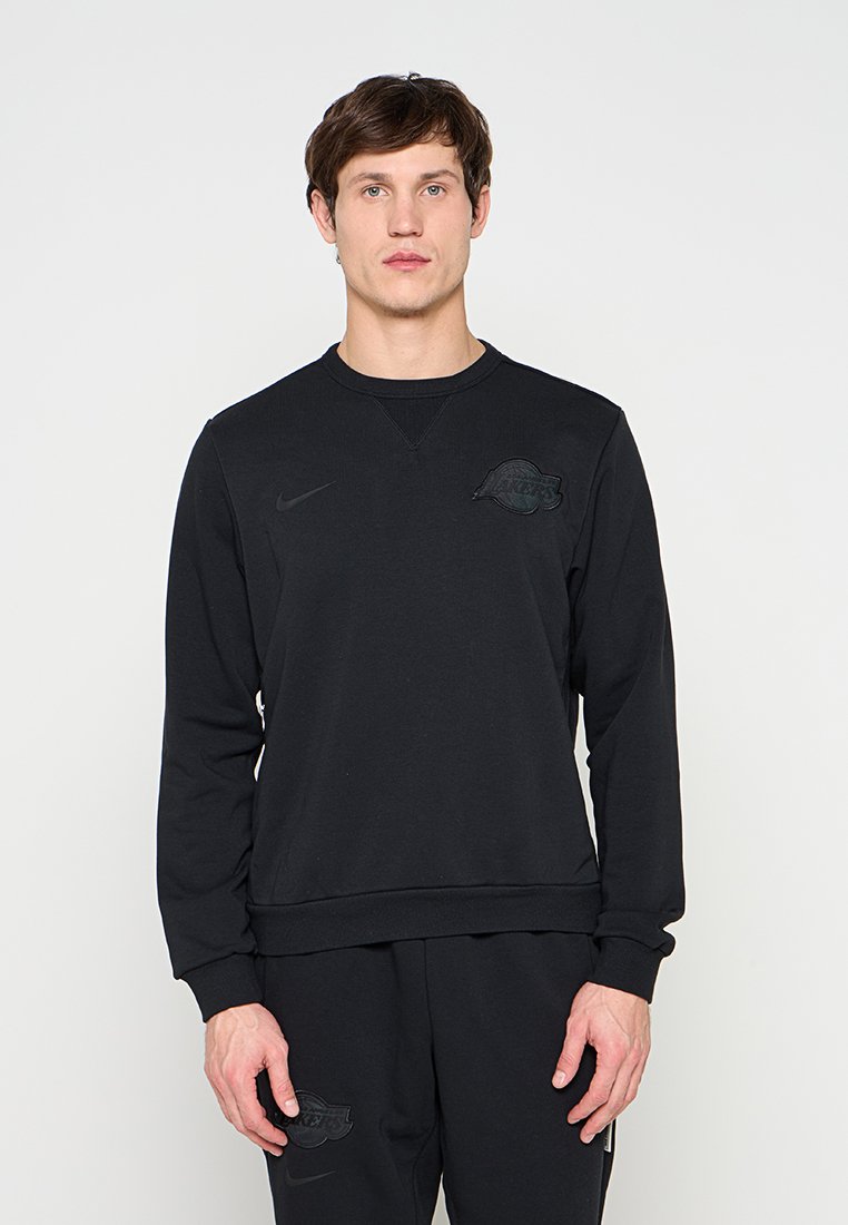 Mann mit schwarzem Langarm-Sweatshirt und passenden schwarzen Hosen, auf Brust und Oberschenkel mit dezenten schwarzen Logos, vor einfarbigem Hintergrund stehend.