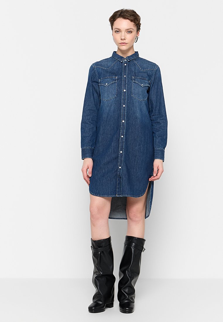 Denimhemdkleid mit langen Ärmeln, Knopfleiste, zwei Brusttaschen und asymmetrischem Saum. Dunkelblaue Farbe, weiche Textur und strukturierte Gestaltung.