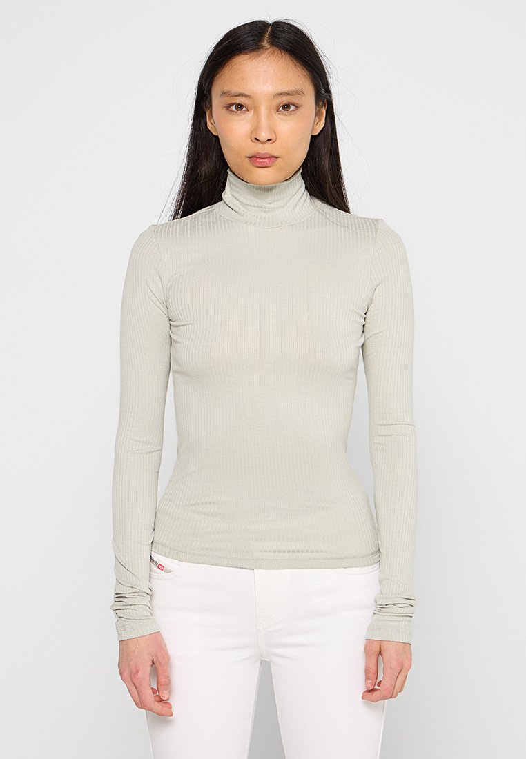 Hellgrauer, gerippter Rollkragenpullover mit langen Ärmeln, der eine taillierte Passform und eine dezente Textur aufweist. Weiße Jeans sind am unteren Ende sichtbar.