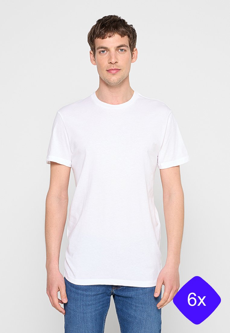 Junger Mann trägt ein schlichtes weißes Kurzarm-T-Shirt und blaue Jeans, steht vor einem neutralen hellen Hintergrund.