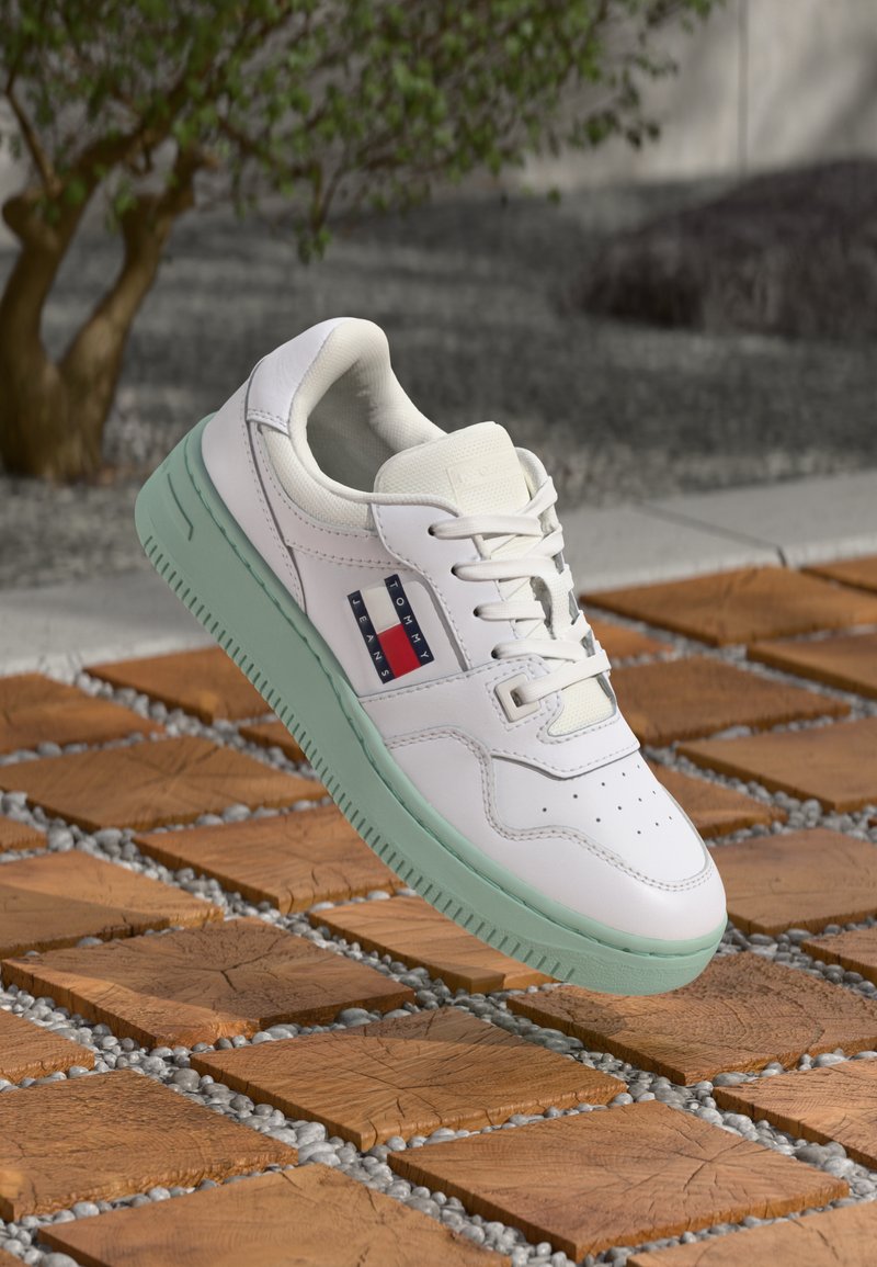 Weiße Sneaker mit grüner Sohle, Obermaterial aus glattem Leder, durchbrochene Zehenpartie und Markenpatch in Rot und Marineblau. Über Holzfliesen erhöht.