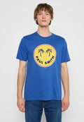 Junger Mann trägt ein blaues T-Shirt mit einem gelben runden Logo, das zwei schwarze Palmen und den Text "PAUL SMITH" zeigt.