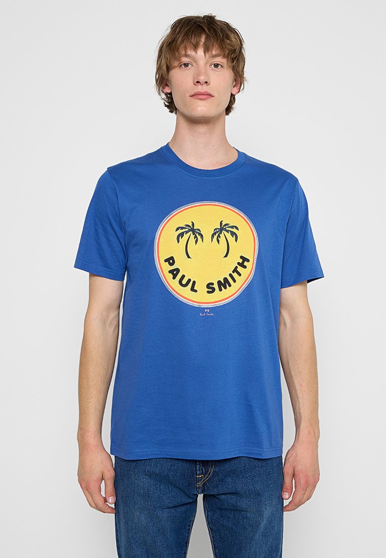 Junger Mann trägt ein blaues T-Shirt mit einem gelben runden Logo, das zwei schwarze Palmen und den Text "PAUL SMITH" zeigt.