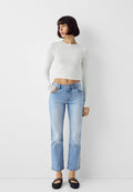 Weißes langärmliges Cropped-Top, hellblaue gerade geschnittene Jeans und schwarze geflochtene Ballerinas. Das Model steht mit einer Hand in der Tasche vor einem neutralen Hintergrund.