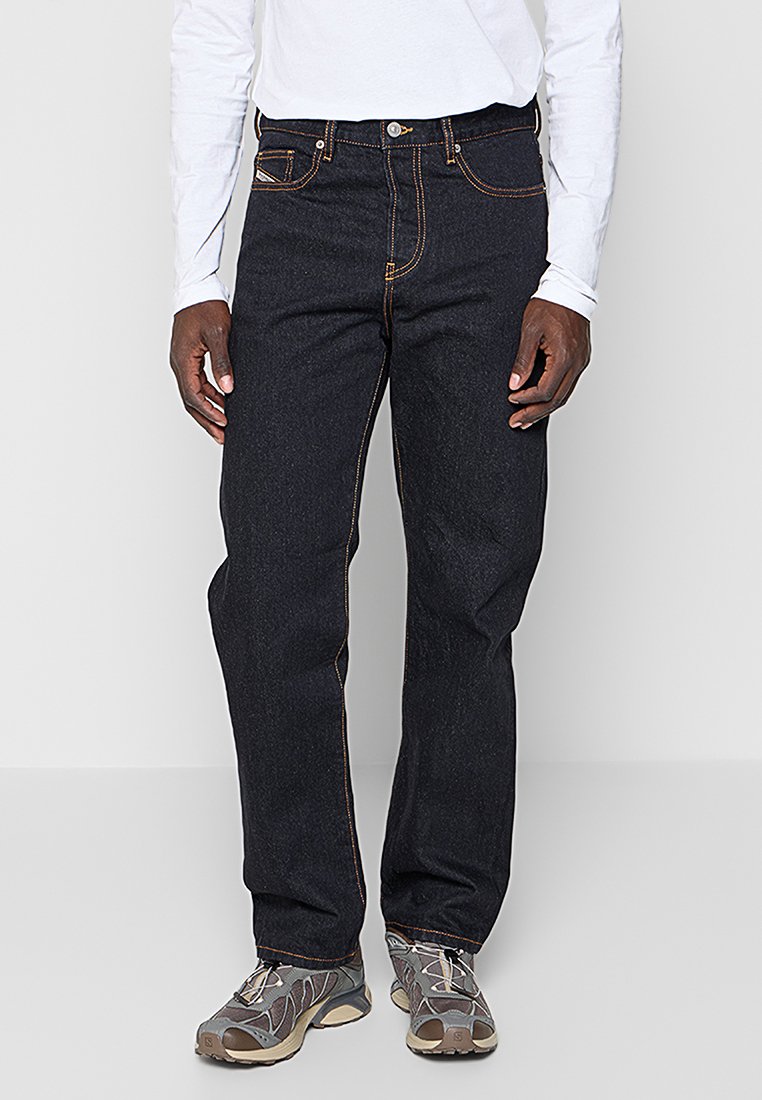 Dunkelblaue Jeans aus Denim mit orangener Naht, gerade geschnitten, fünf Taschen und silbernen Beschlägen, kombiniert mit grauen Turnschuhen.