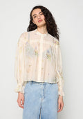 Frau mit lockigem dunklem Haar, die eine durchsichtige weiße Blumen-Bluse mit Knöpfen und hellblaue, hoch taillierte Jeans trägt, steht vor einem weißen Hintergrund.