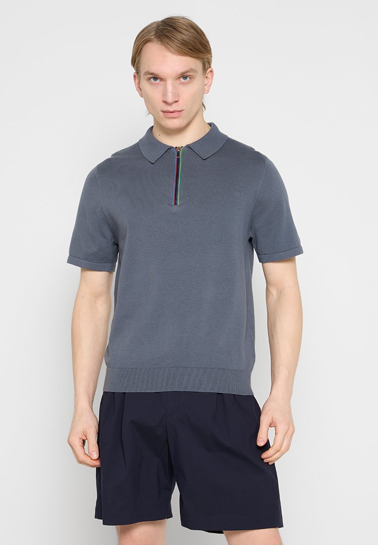Graues kurzärmeliges Poloshirt mit Reißverschluss am Ausschnitt und farbenfrohen Details. Gerippte Textur und figurbetonter Saum. Getragen zu navyblauen Shorts.