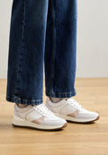 Weiße Sneaker mit beigen Akzenten, glatten und strukturierten Materialien, runder Zehenform und Schnürdesign, kombiniert mit dunkelblauen, weit geschnittenen Jeans.