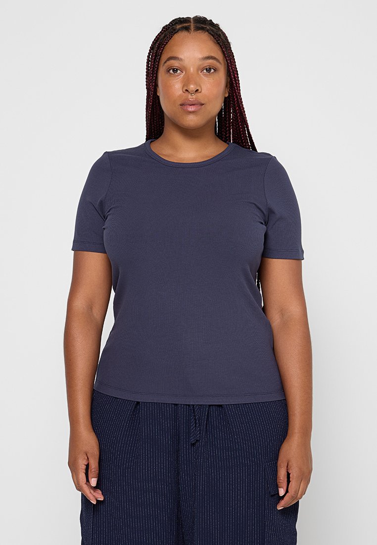 Frau mit langen geflochtenen Haaren, die ein enges dunkelblaues kurzärmliges Shirt und dunkelblau gestreifte Hosen trägt, steht vor einem schlichten Hintergrund.