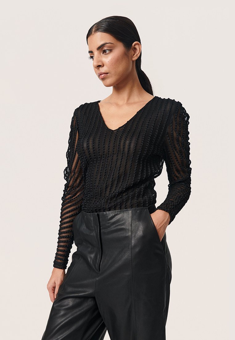 Schwarzes, transparentes Langarmshirt mit strukturierten vertikalen Streifen, einem V-Ausschnitt und figurschmeichelndem Schnitt, kombiniert mit hochgeschnittenen schwarzen Lederhosen.