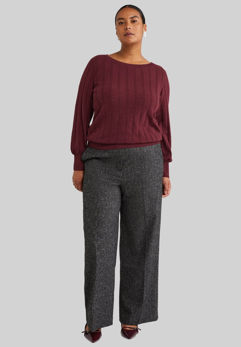 Burgunderfarbener Strickpullover mit gerippter Textur, kombiniert mit grauen weit geschnittenen Hosen, die ein dezentes gesprenkeltes Muster aufweisen. Braune Schuhe mit Akzenten.