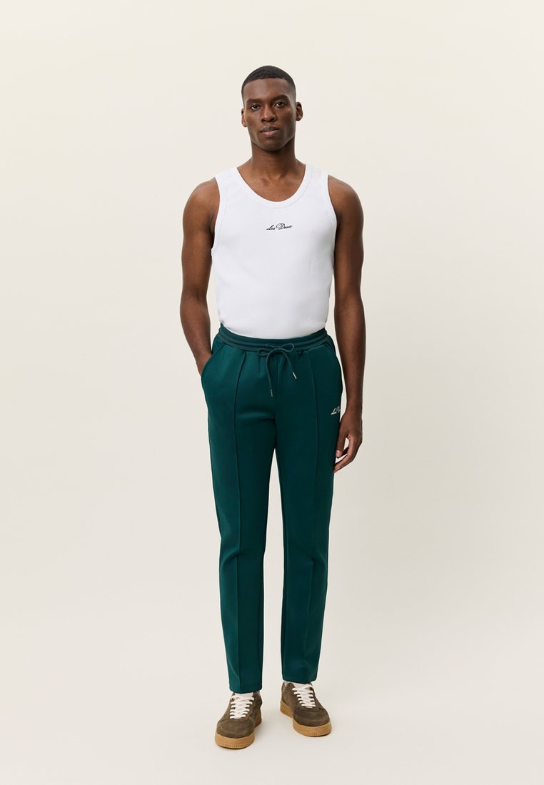 Grüne Jogginghose mit elastischem Bund und Kordelzug, kombiniert mit einem weißen ärmellosen Oberteil. Strukturierter Stoff und figurbetontes Design. Braune Sneakers.