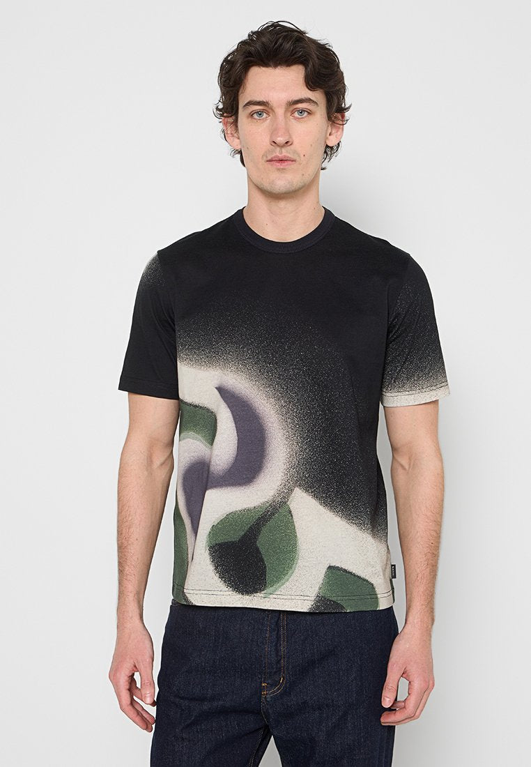 Mann trägt ein schwarzes und beiges T-Shirt mit Farbverlauf und abstrakten grünen, grauen und schwarzen Formen, kombiniert mit dunkelblauen Jeans, steht vor einem einfarbigen Hintergrund.