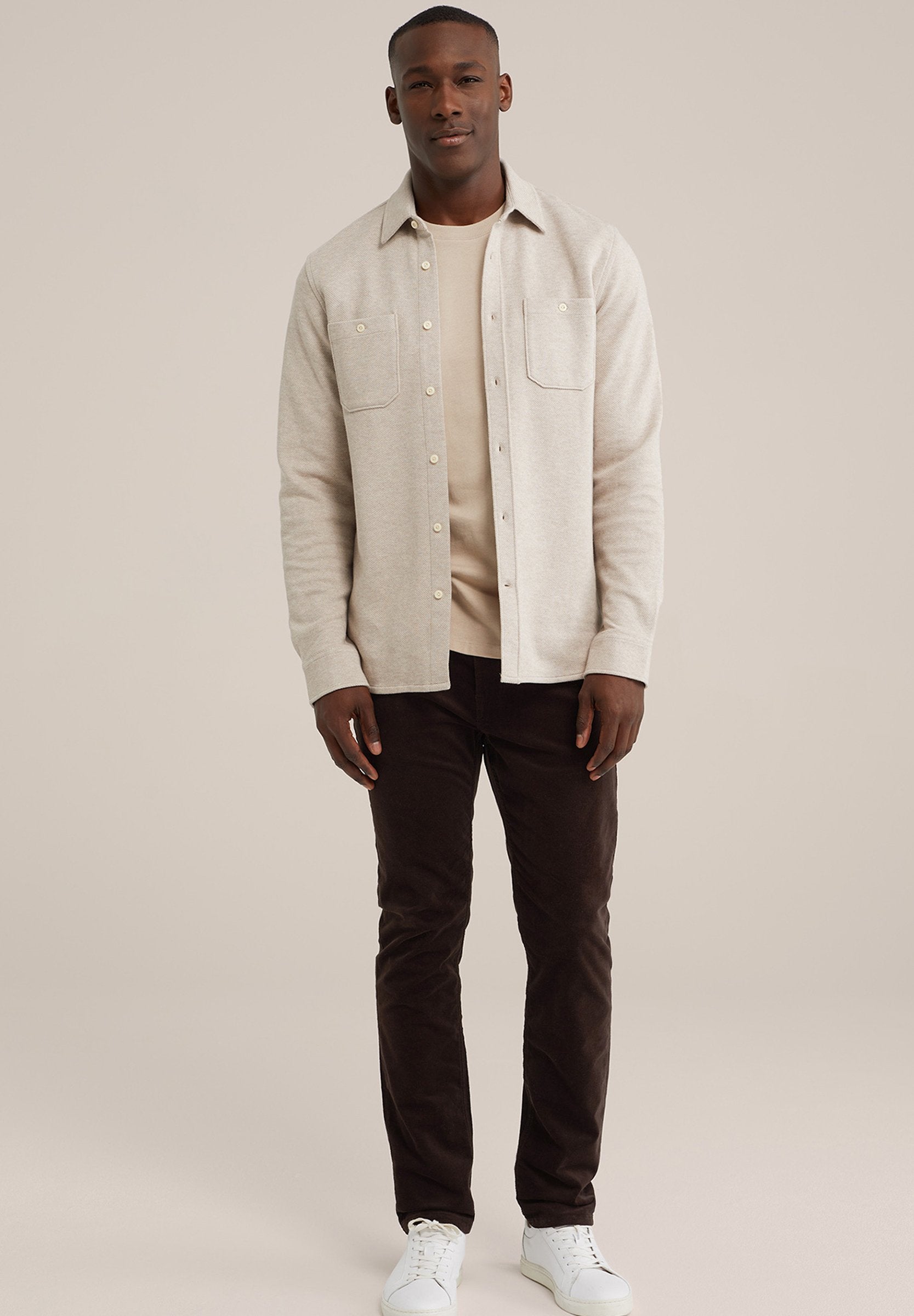 Helle beige Knopfleiste mit zwei Fronttaschen, über einem cremefarbenen Oberteil geschichtet. Dunkelbraune Hose und weiße Turnschuhe runden das Outfit ab.