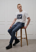 Weißes T-Shirt mit blauem Grafikdruck und "HUGO"-Schriftzug, kombiniert mit blau gemusterten Hosen. Schwarze klobige Schuhe, sitzend auf einem hölzernen Hocker.