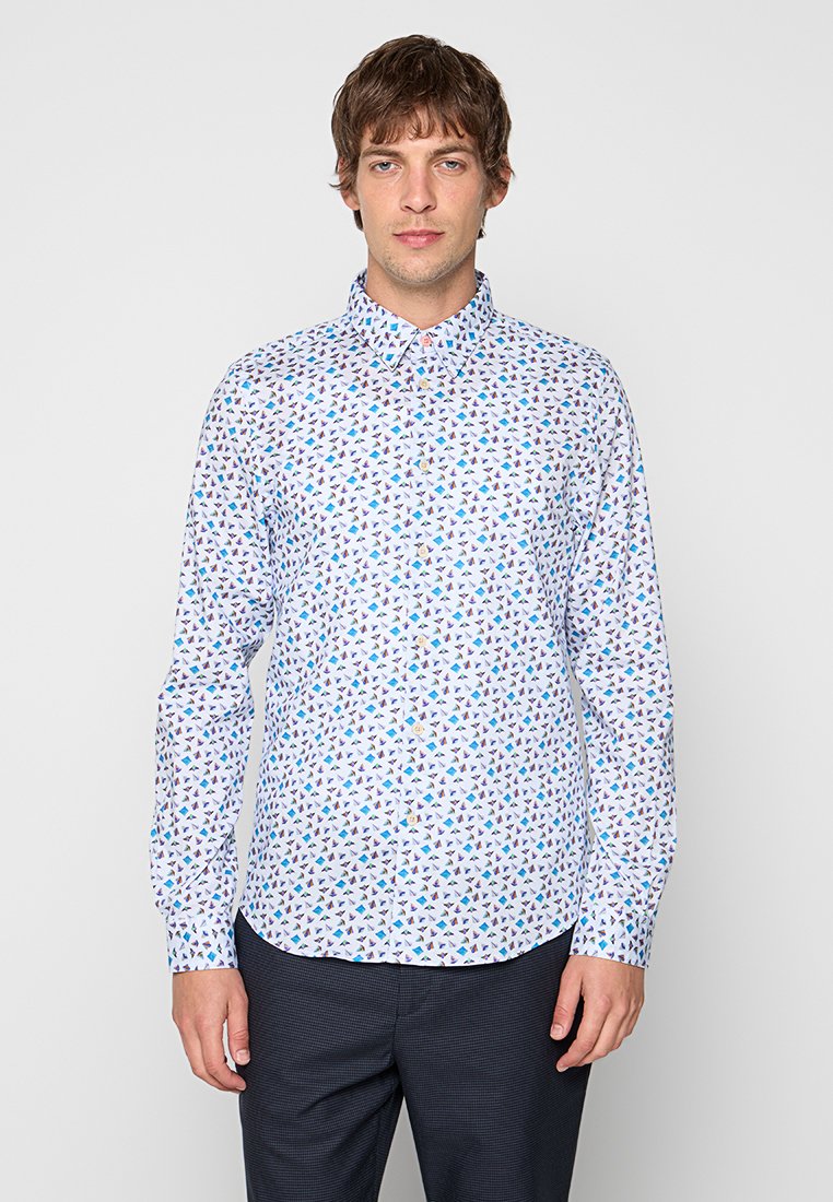 Mann trägt ein tailliertes weißes Hemd mit blauem und lila geometrischem Muster und dunkle Hose, steht vor einem schlichten Hintergrund.