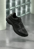 Schwarzer Sportschuh mit Mesh- und Leder-Details, schwebt über einem polierten Betonboden in der Nähe einer Glassteinwand in einem minimalistischen Innenraum.