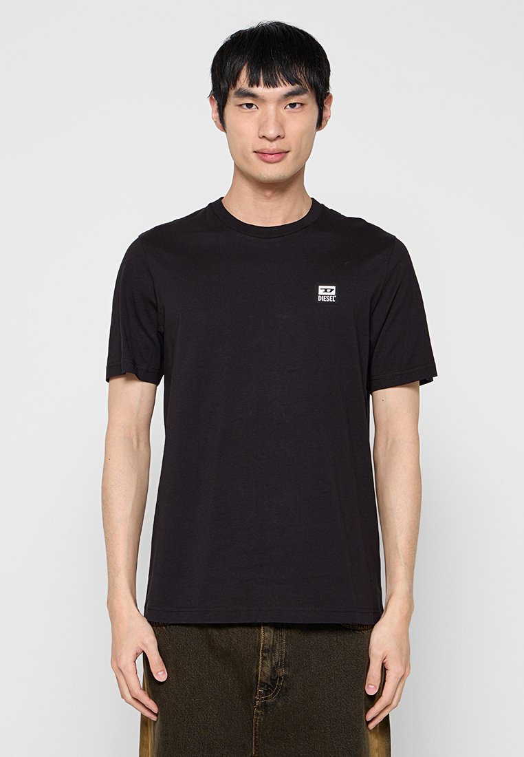 Schwarzes Baumwoll-T-Shirt mit Rundhalsausschnitt und kurzen Ärmeln, versehen mit einem kleinen weißen rechteckigen Etikett mit der Aufschrift "DIESET" auf der Brust.