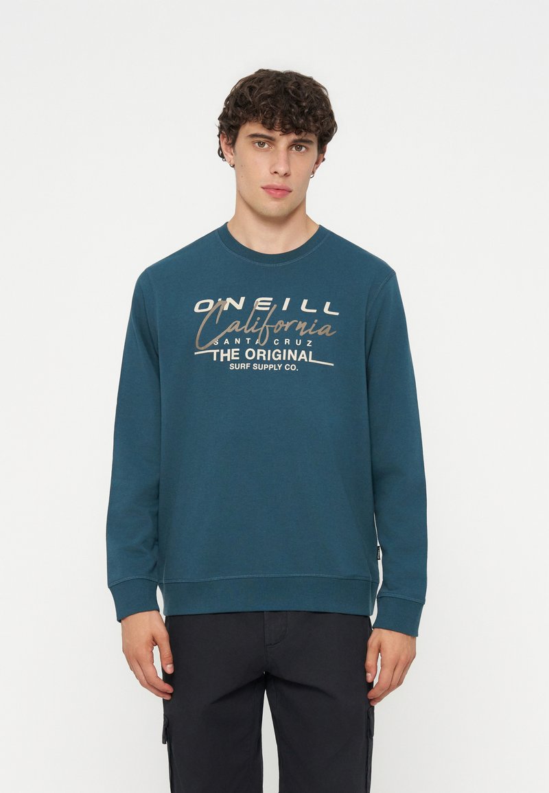 Junger Mann mit dunklem lockigem Haar, der ein türkisfarbenes Sweatshirt mit beigem Schriftzug „O'Neill California Santa Cruz Surf Supply Co.“ trägt.