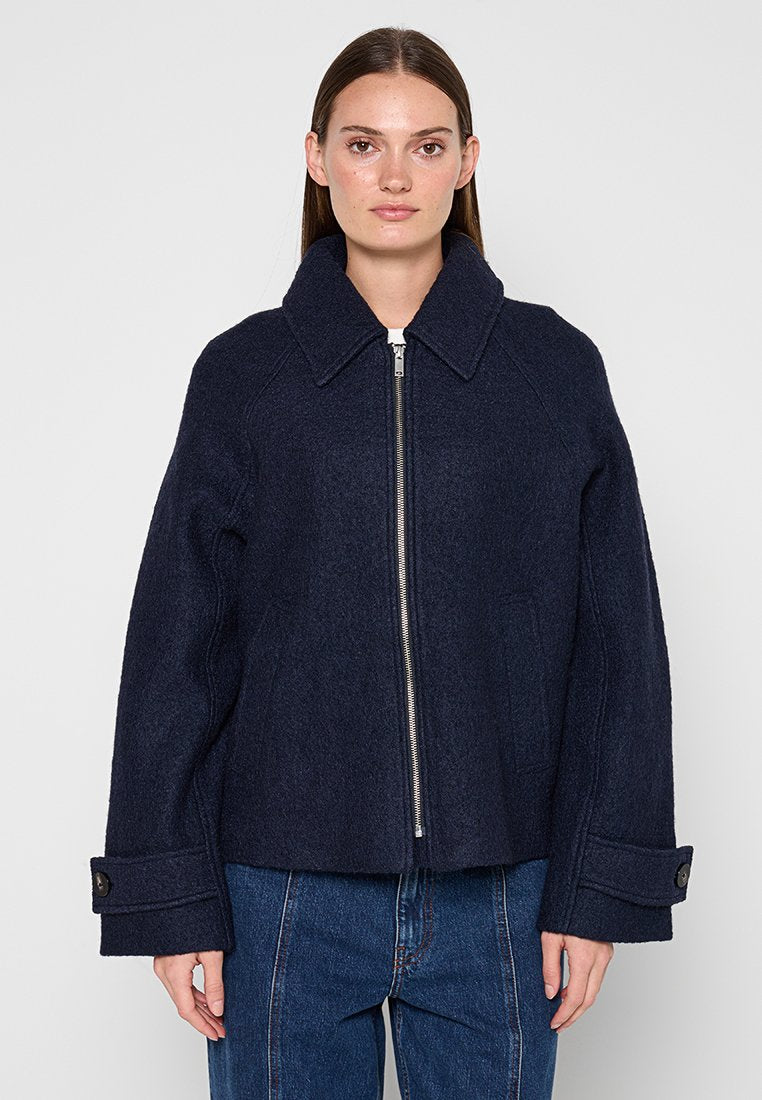 Frau trägt eine dunkelmarineblaue Wolljacke mit Reißverschluss, breitem Kragen und Manschettenriemen, kombiniert mit blauen Jeans, stehend vor einfarbigem Hintergrund.