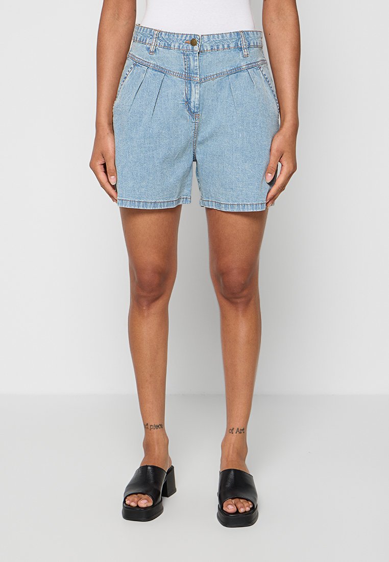 Frau trägt hellblaue plissierte Denim-Shorts und schwarze offene Sandalen mit Blockabsatz, steht vor einem einfarbigen hellen Hintergrund.