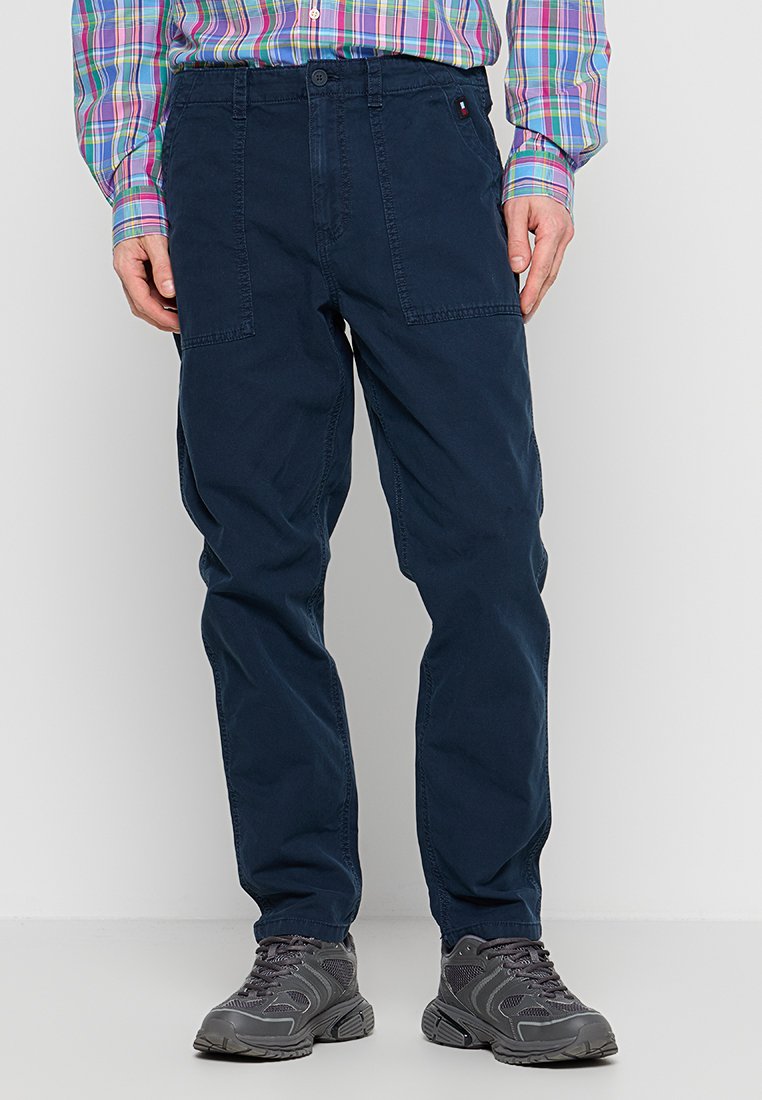 Marineblaue Baumwollhosen mit geradem Schnitt, zwei Vordertaschen und Knopfverschluss. Kombiniert mit grauen Sneakers.